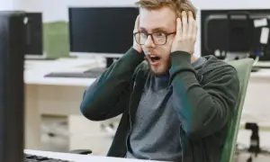 Un homme choqué, les mains sur les oreilles, devant son écran. Il semble avoir découvert un problème majeur dans le système d'information ou le système informatique.