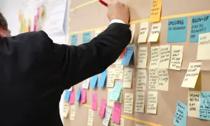 Un homme en costume noir écrit sur un tableau rempli de post-its, planifiant un exemple de projet informatique.