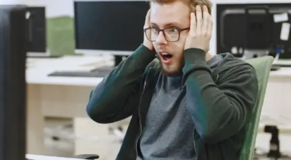 Un homme choqué, les mains sur les oreilles, devant son écran. Il semble avoir découvert un problème majeur dans le système d'information ou le système informatique.