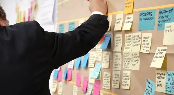 Un homme en costume noir écrit sur un tableau rempli de post-its, planifiant un exemple de projet informatique.