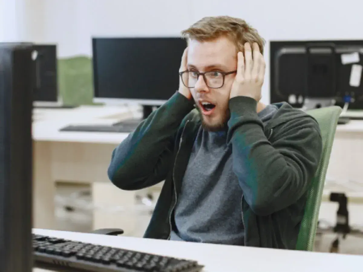 Un homme choqué, les mains sur les oreilles, devant son écran. Il semble avoir découvert un problème majeur dans le système d'information ou le système informatique.