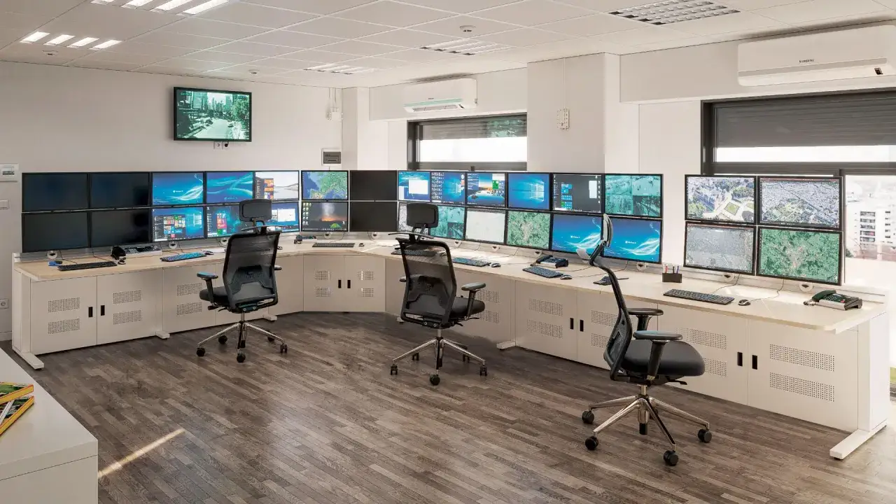 Salle de contr&ocirc;le avec de nombreux &eacute;crans affichant des cartes et des flux vid&eacute;o, g&eacute;r&eacute;e par un logiciel de supervision. Des chaises de bureau ergonomiques sont dispos&eacute;es devant le poste de travail.