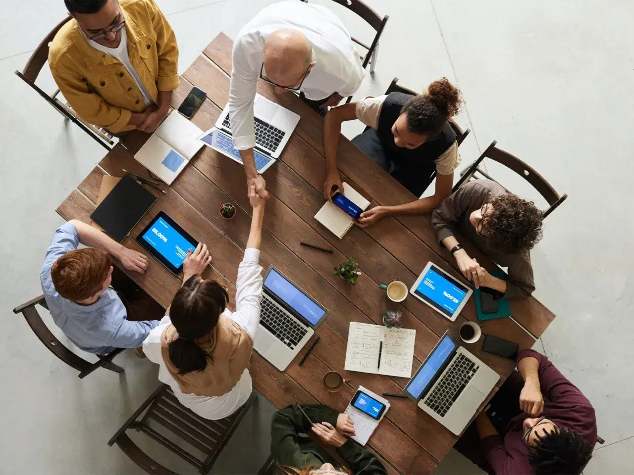 Une équipe diverse se réunit autour d'une table en bois, échangeant des idées et des poignées de main. Une scène qui illustre parfaitement le travail d'équipe et la collaboration.