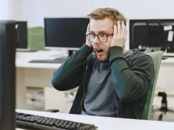 Un homme choqué, les mains sur les oreilles, devant son écran. Il semble avoir découvert un problème majeur dans le système d'information ou le système informatique.