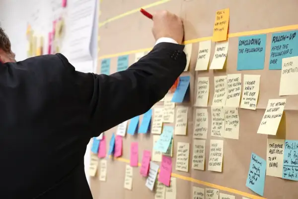Un homme en costume noir écrit sur un tableau rempli de post-its, planifiant un exemple de projet informatique.
